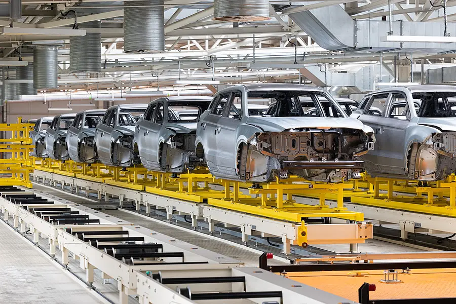 Two rows of silver partially-constructed cars on yellow and white conveyer belts in a car factory. Crumple zones 