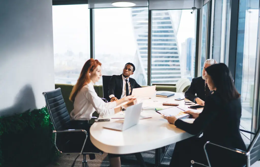 Group of multiracial colleagues in formal suits gathering at table in modern workspace with panoramic windows and discussing project details