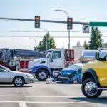 This image shows a truck accident investigation scene in Scranton, Pennsylvania, handled by Munley Law Personal Injury Attorneys, reachable at (570) 865-4699. The photo captures an intersection with clear traffic signals, an 18-wheeler involved in a collision with damaged passenger vehicles, and emergency vehicles present. The accident appears severe, with a blue car showing heavy front-end damage. Munley Law offers professional legal representation in truck accident cases, assisting Scranton residents affected by similar incidents.