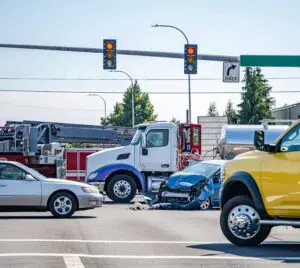 This image shows a truck accident investigation scene in Scranton, Pennsylvania, handled by Munley Law Personal Injury Attorneys, reachable at (570) 865-4699. The photo captures an intersection with clear traffic signals, an 18-wheeler involved in a collision with damaged passenger vehicles, and emergency vehicles present. The accident appears severe, with a blue car showing heavy front-end damage. Munley Law offers professional legal representation in truck accident cases, assisting Scranton residents affected by similar incidents.