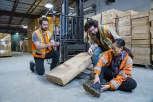 Warehouse workers in Allentown assisting an injured employee seated on the floor near a forklift, illustrating a typical workplace injury scenario. Munley Law's workers' compensation attorneys in Allentown, PA, available at (855) 866-5529, offer representation on a contingency fee basis, charging no upfront costs. The injured employee holds their ankle, clearly indicating pain from a workplace accident involving warehouse equipment. Colleagues provide immediate support, highlighting the necessity of skilled legal guidance after workplace injuries. Munley Law helps clients navigate workers' compensation claims effectively, ensuring clear, upfront attorney fees.
