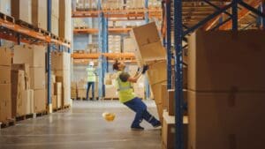 A worker in a high-visibility vest is about to be struck by falling boxes in a warehouse. The hard hat has already fallen, indicating a workplace safety hazard. This image represents a typical case handled by a Philadelphia workers compensation lawyer at Munley Law. Call 215-515-7747 for help filing a workers’ compensation claim in Pennsylvania.