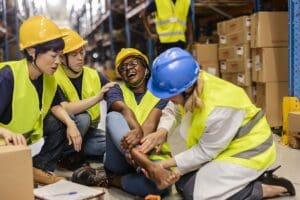An injured warehouse worker sits on the warehouse floor holding her ankle in pain, surrounded by concerned coworkers who provide immediate first aid. Munley Law offers experienced legal representation for Pennsylvania workers injured on the job, helping secure medical coverage and wage loss benefits. With offices in Scranton, Allentown, and throughout Pennsylvania, Munley Law attorneys are available 24/7 at844-686-5397 to navigate workers' compensation claims. This image underscores the importance of prompt medical and legal support following workplace injuries.